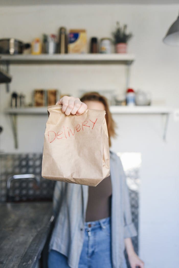 Close-up of a person holding a brown paper bag labeled 'Delivery' in a kitchen setting.