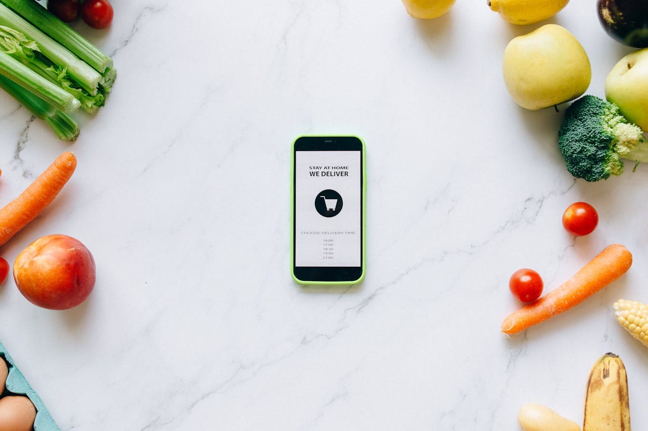 Mobile delivery app surrounded by fresh fruits and vegetables on a marble countertop.