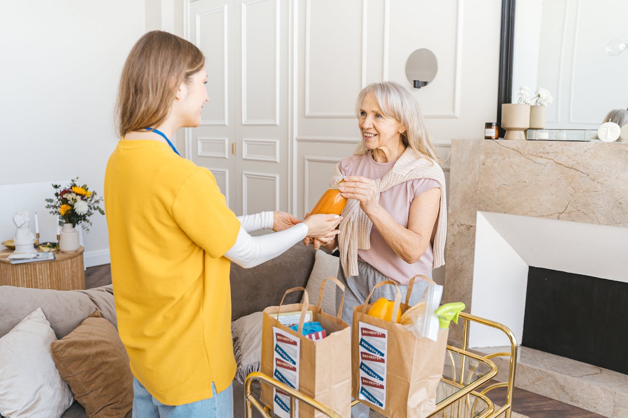 A senior woman receiving groceries from a young caregiver in a cozy living room setting.
