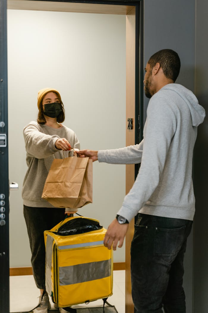 services-03 A delivery person hands food to a masked customer at an apartment door, showcasing a contactless exchange.