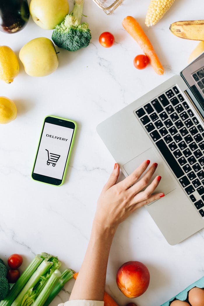 A top view of online grocery shopping using a laptop and smartphone surrounded by fresh vegetables and fruits.